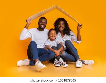 Housing For Young Family Concept. Young Black Father, Mother And Daughter Sitting Under Symbolic Roof Dreaming Of New Home Over Yellow Background