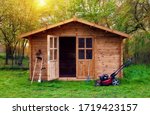 Hovel after work in evening, golden hour. Garden shed (front view) with hoe, string trimmer,  rake and grass-cutter. Gardening tools shed. Garden house on lawn in the sunset. Wooden tool-shed. 