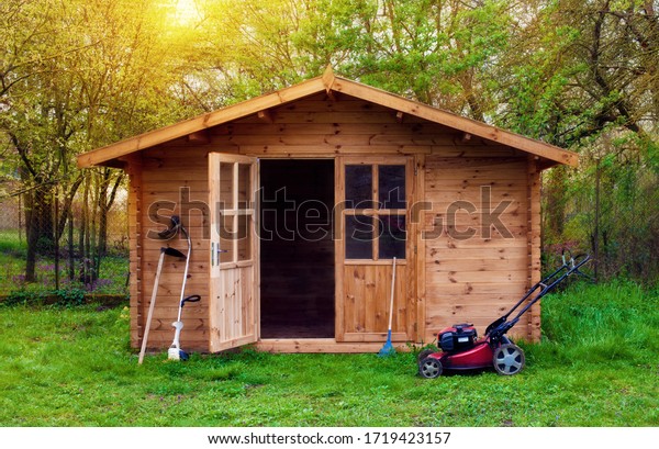 Hovel after work in evening, golden hour. Garden shed\
(front view) with hoe, string trimmer,  rake and grass-cutter.\
Gardening tools shed. Garden house on lawn in the sunset. Wooden\
tool-shed. 