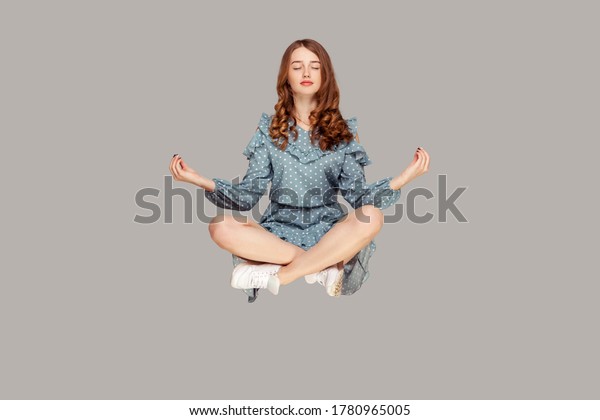 Hovering in air. Calm peaceful relaxed girl\
ruffle dress levitating with mudra gesture hands up, closed eyes,\
meditating sitting in yoga position. indoor studio shot isolated on\
gray background