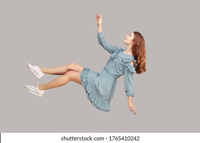 Hovering in air. Cheerful smiling pretty girl in vintage ruffle dress levitating flying in mid-air, looking up happy dreamy and raising hand to catch. indoor studio shot isolated on gray background