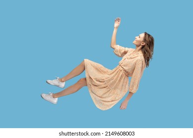 Hovering in air. Cheerful smiling pretty girl in yellow dress levitating flying in mid-air, looking up happy dreamy and raising hand to catch. indoor studio shot isolated on blue background