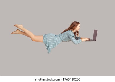 Hovering in air. Happy cheerful girl ruffle dress levitating with laptop, typing keyboard, reading good news message on computer while flying in mid-air. studio shot isolated on gray background