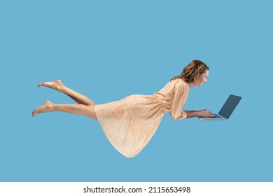 Hovering in air. Happy cheerful girl in yellow dress levitating with laptop, typing keyboard, reading good news message on computer while flying in mid-air. studio shot isolated on blue background