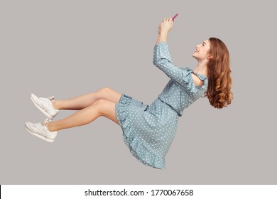 Hovering in air. Smiling girl ruffle dress levitating with mobile phone, reading message chatting happy joyful in social network online, surfing web while flying. indoor studio shot isolated on gray