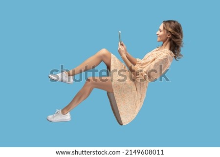 Hovering in air. Smiling girl in yellow dress levitating with mobile phone, reading message chatting happy joyful in social network online, surfing web while flying. indoor shot isolated on blue