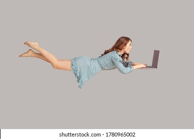Hovering in air. Surprised excited girl ruffle dress levitating with laptop, typing keyboard, reading shocking news message on computer while flying in mid-air. studio shot isolated on gray background