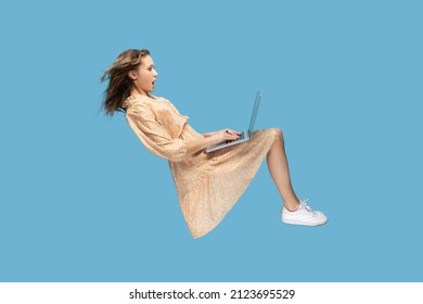 Hovering in air. Surprised girl in yellow dress levitating, looking at laptop screen shocked amazed, surfing web social networks while flying in mid-air. indoor studio shot isolated on blue background