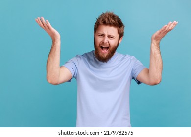 How could you? Portrait of frustrated handsome bearded man standing with raised hands and indignant face asking why, shrugging shoulders. Indoor studio shot isolated on blue background.