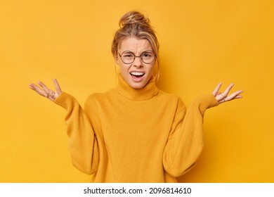How could you. Annoyed indignant young woman spreads palms smirks face looks angrily at camera quarrels with someone feels puzzled wears spectacles and sweater isolated over yellow background - Shutterstock ID 2096814610