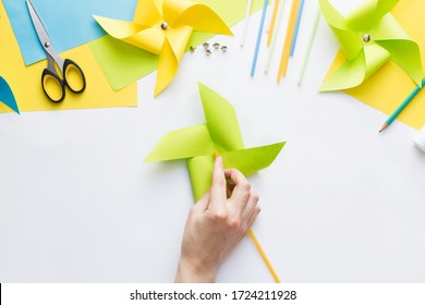 How To Make Paper Green Windmill Toy With Children At Home. Step By Step Instructions. Hands Making DIY Summer Project. Top View. Step 11. Turn Pinwheel To Other Side And Attach Straw To Push Pin