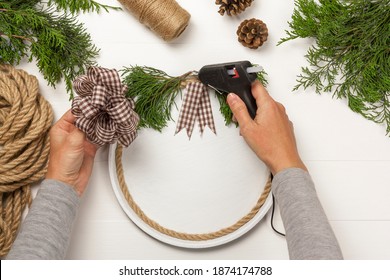 How To Make Pizza Pan Christmas Door Hanger Sign. Step-by-step Instruction. Step 4. Female Hands Glue Fir Branches And Cotton Ribbon Bow To Pizza Pan With Hot Glue Gun. DIY Craft Farmhouse Decor.