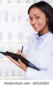 How May I Help You? Confident Young African Woman In Lab Coat Making Notes In Clipboard And Smiling To You While Standing In Drugstore 