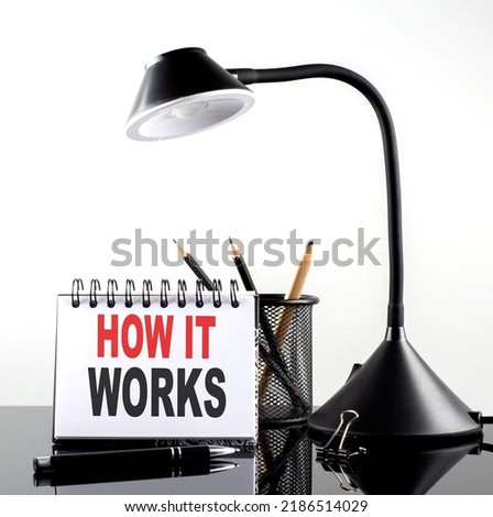 HOW IT WORKS text on notebook with pen and table lamp onblack background