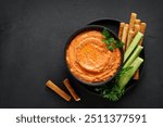 Htipiti - Greek red pepper and feta dip in black bowl with bread sticks and cucumbers, on dark background. top view, copy space