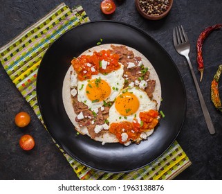 Huevos Rancheros, Mexican Fried Egg On A Wheat Tortilla With Tomato Salsa, Bean Paste And Feta Cheese On A Black Plate On A Black Concrete Background. Mexican Food.