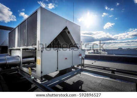A huge air conditioning unit on the roof of the building. In the background of blue sky with shining sun. Focus is at the front of the air conditioner, the other parts of image slightly blurred.
