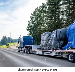 Huge Blue Classic Big Rig Industrial Semi Truck Transporting Covered With Tarp Heavy Commercial Cargo On Step Down Semi Trailer Running On The Wide Highway Road With Trees On The Side