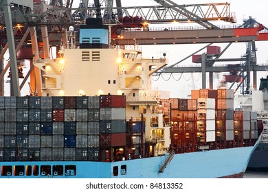 A Huge Container Ship Being Unloaded At Dawn In An Industrial Harbor By Overhead Cranes