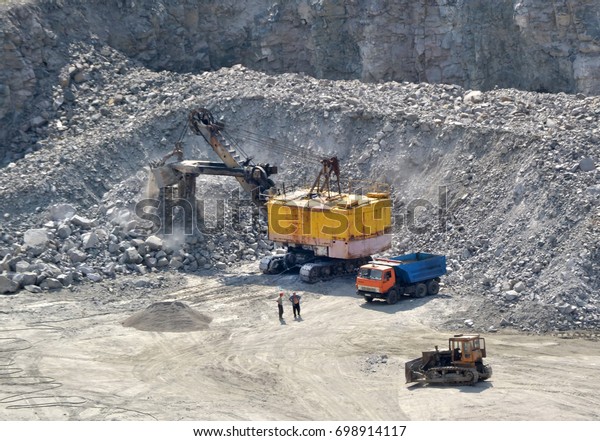 Huge excavator,\
truck and man standing next on granite quarry. Yellow carver\
machine, excavator, stonecutter for industrial granite stands in a\
quarry near granite stones
