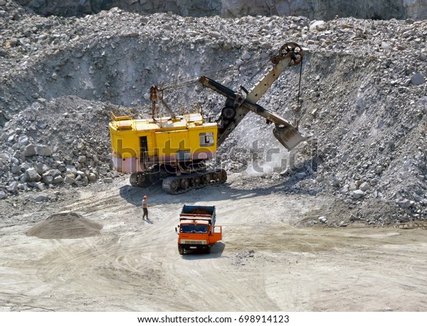 Huge excavator,\
truck and man standing next on granite quarry. Yellow carver\
machine, excavator, stonecutter for industrial granite stands in a\
quarry near granite stones