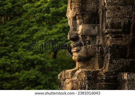 A huge face sculpted in stone, watches over visitors to the temple of Bayon, Angkor, Cambodia, in the background, a wall of green jungle surrounds it