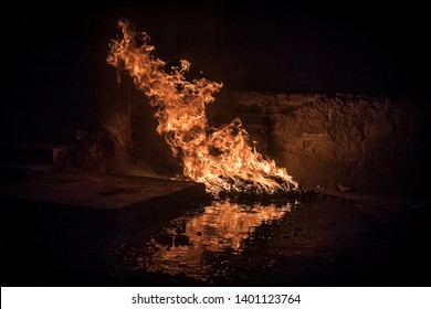 Huge Fire Burning Off Of Lubricant Oil And Motor Parts Beside A Garage, The Reflection Of The Flames Can Be Seen In The Oil Spill On The Ground