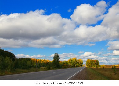 Huge fluffy clouds float on the blue sky above the road, fields and trees. Autumn trees, on the field and on the road there are bizarre shadows. Light and shadow on the autumn day decided to compete