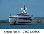 A huge, innovative white yacht is anchored off the coast of Sardinia. The mega yacht rests on blue water, with the mountainous relief and blue sky in the background.