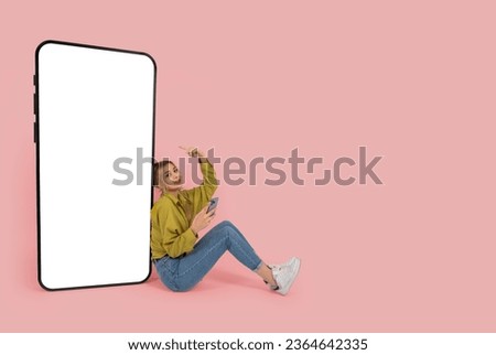 Huge mobile phone mock up, full body view young caucasian blonde girl sit near big huge mobile phone mock up. Holding smartphone, pointing empty blank screen phone. Copy space, advertising concept.