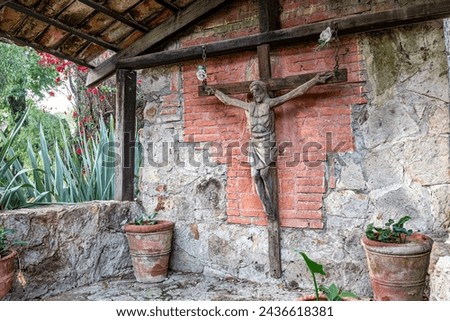Huge old wooden cross with crucified Christ in shed of ranch, tile roof, worn brick walls, clay pots with small green plants, red bougainvillea and trees in background, sunny day in Jalisco Mexico