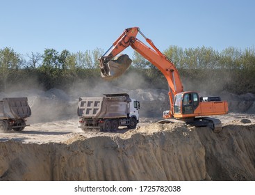 huge orange crawler excavator and
 two gray construction dump trucks in the process of working on a sand pit against a blue sky. sunny day. Dust loading sand