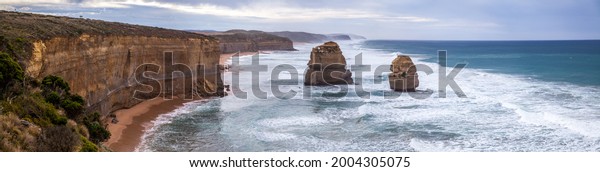 A huge panorama of orange steep cliffs and rocky islands\
in the ocean near Twelve Apostles Sea Rocks near Great Ocean Road,\
Port Campbell National Park, Australia. Wide waves rolling to the\
seashore. 