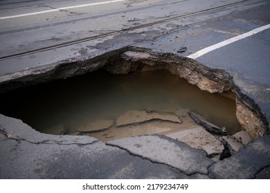 Huge Sinkhole On Busy Asphalt Road Surface On Which Cars Drive. Accident Situation On A City Street Due To Cracks In Asphalt. Broken Hole Filled With Muddy Water.