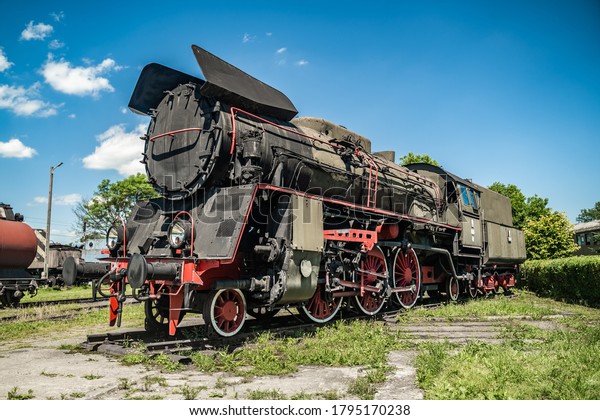 Huge vintage steam locomotive, black and red\
painted steel train. Coal-powered steam express stands on tracks.\
Classic gigantic heavy railway machinery. Side view of power parts\
of machine.