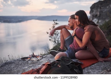 Hugging couple with backpack sitting near the fire on top of mountain enjoying view coast a river or lake. Traveling along mountains and coast, freedom and active lifestyle concept.