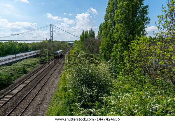 Hull, UK - 12 May 2019: The train track from\
Hull train station alongside Clive Sullivan Way motorway near Hull\
city centre with yellow train and the Humber Bridge single span\
bridge in the background