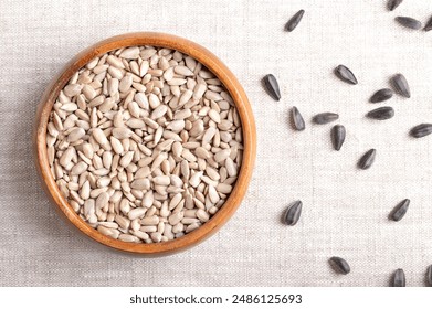 Hulled sunflower seeds in a wooden bowl on linen fabric. Fruits of the oilseed Helianthus Annuus, the common sunflower. On the right side whole seeds with black hulls. Close-up from above, food photo. - Powered by Shutterstock