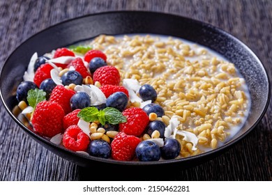 Hulled Whole-grain Oat Porridge Cooked With Coconut Milk Topped With Fresh Raspberries, Blueberries And Coconut Chips In Black Bowl On Wood Table, Horizontal View From Above