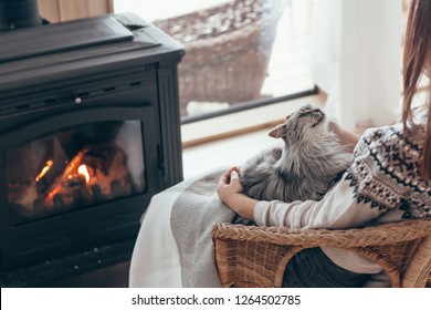 Human With Cat Relaxing In Wicker Armchair By The Fire Place In Wooden Cabin. Warm And Cozy Winter Holiday Concept.