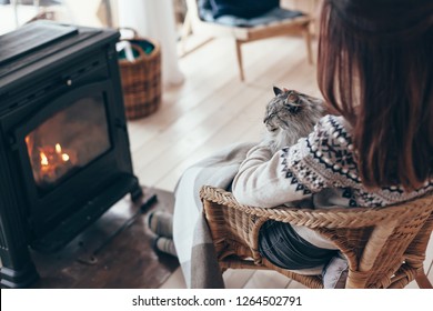 Human With Cat Relaxing In Wicker Armchair By The Fire Place In Wooden Cabin. Warm And Cozy Winter Holiday Concept.