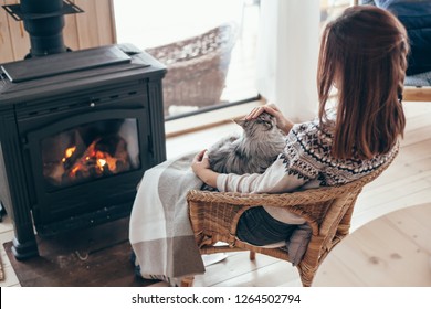 Human With Cat Relaxing In Wicker Armchair By The Fire Place In Wooden Cabin. Warm And Cozy Winter Holiday Concept.