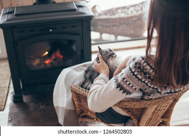 Human With Cat Relaxing In Wicker Armchair By The Fire Place In Wooden Cabin. Warm And Cozy Winter Holiday Concept.