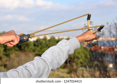 Human (child) Is Throwing Stone With Slingshot - Wooden Slingshot With A Stretched Rubber Band. Slingshot Is Ready To Fire