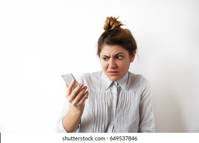 Human Emotions, Feelings And Reactions. Attractive Young Woman With Hair Bun Looking At Mobile Phone Seeing Bad News Photos Having Scared Or Shocked Facial Expression, Raising Eyebrows