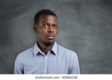 Human Face Expressions And Emotions. Close Up Shot Of Doubtful African Man Dressed Casually Looking Confused And Puzzled, Raising His Brow In Surprise And Indignation, Standing At Blank Blackboard