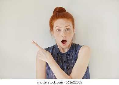 Human face expressions, emotions and feelings. Astonished and shocked young redhead female in sleeveless shirt pointing at blank studio wall, surprised with sale prices, keeping her mouth wide open