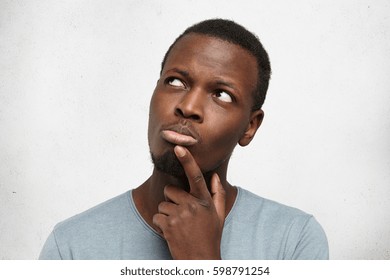 Human face expressions, emotions and feelings. Handsome young African American man looking up with thoughtful and skeptical expression, holding finger on his chin, trying to remember something