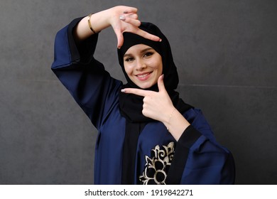 Human face expressions and emotions Positive young beautiful female with Hijab hair cover in Abaya clothing making a square with her fingers, looking at camera and pleasantly smiling