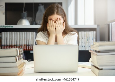 Human face expressions and emotions. Young Caucasian student with long hair covering her face with hands, crying of fatigue, fed up with preparing for exams sitting at the table in front of laptop